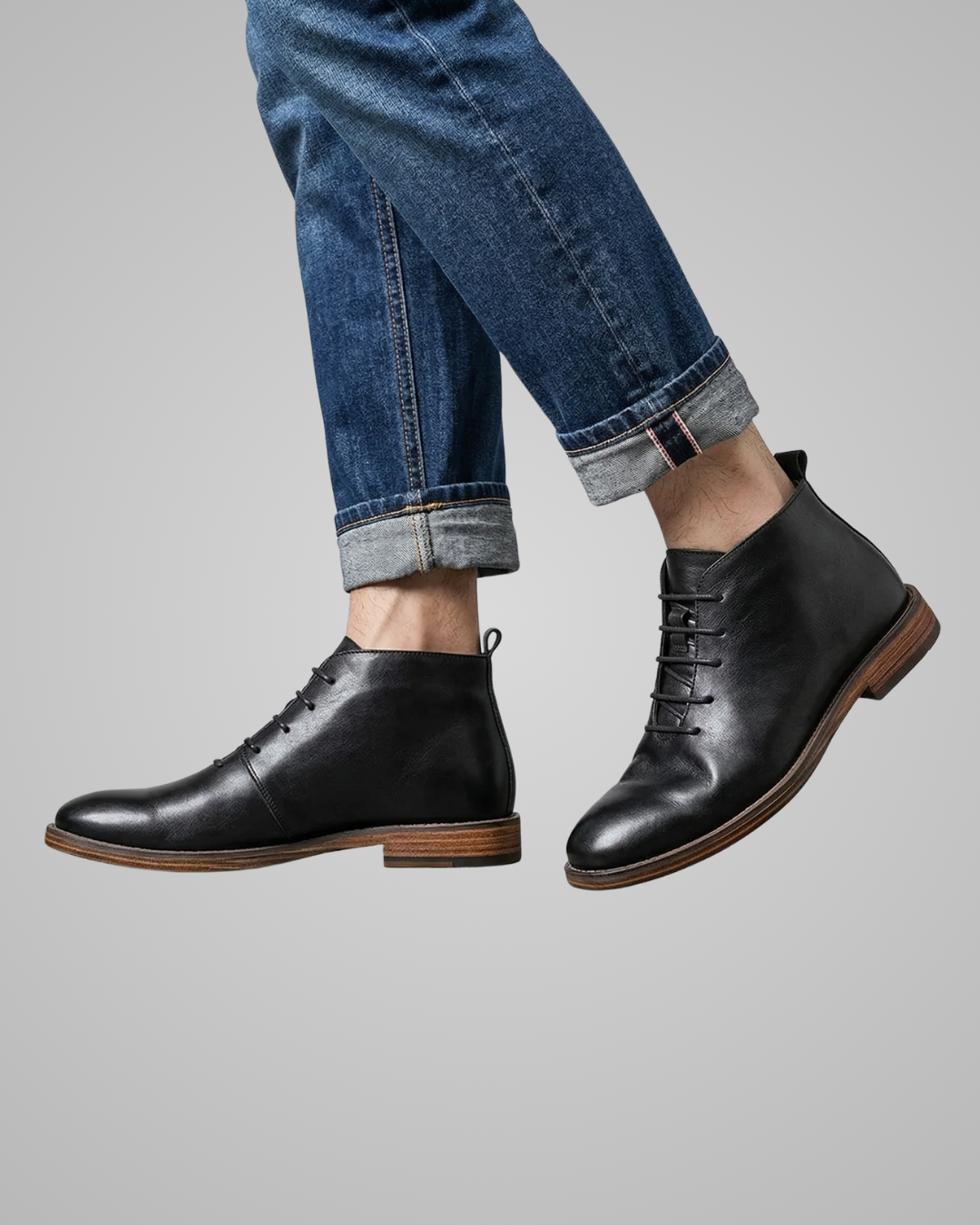 Black leather boots worn with rolled-up blue jeans on a gray background