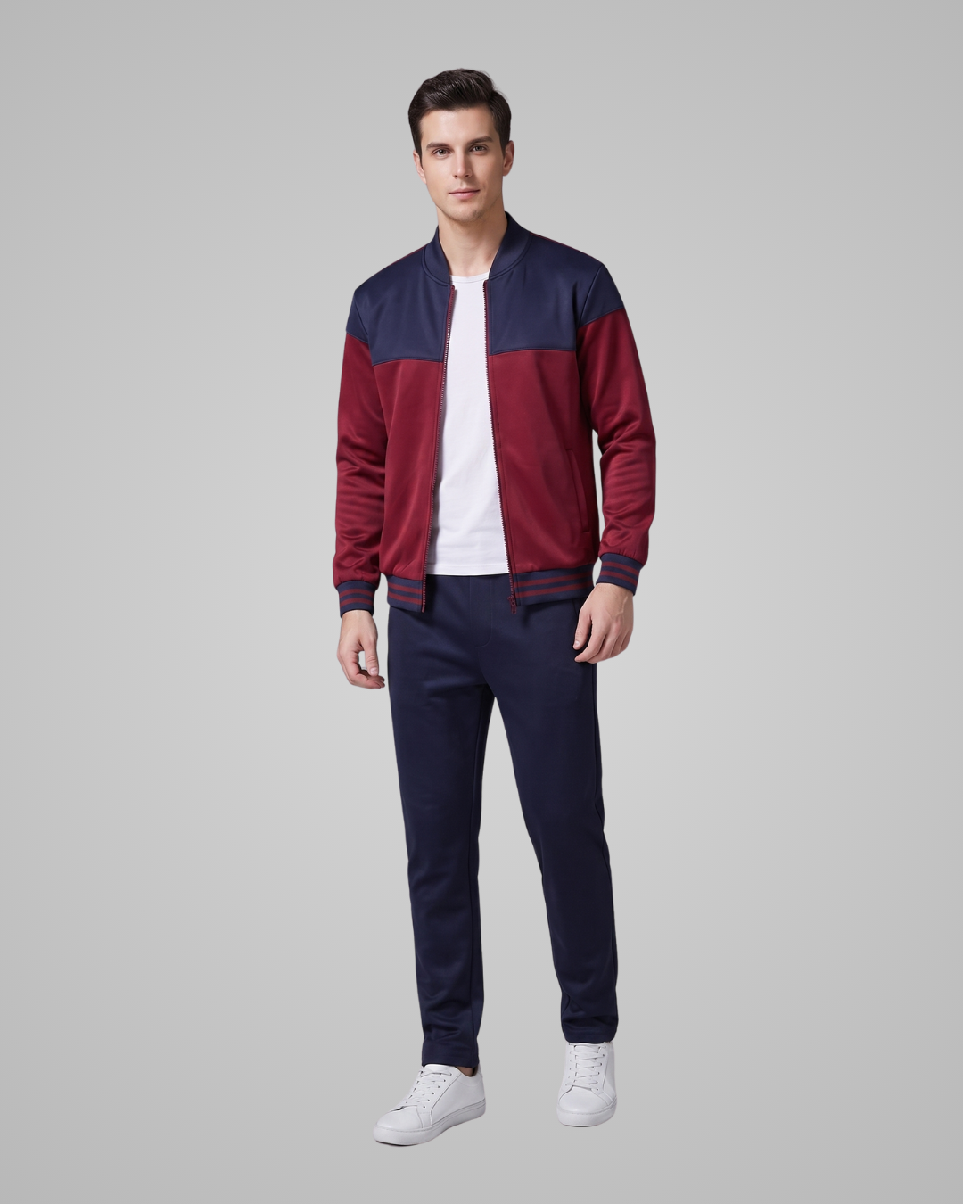A man modeling a color-blocked bomber jacket with navy blue and burgundy panels, paired with navy pants and white sneakers, against a solid gray studio background.