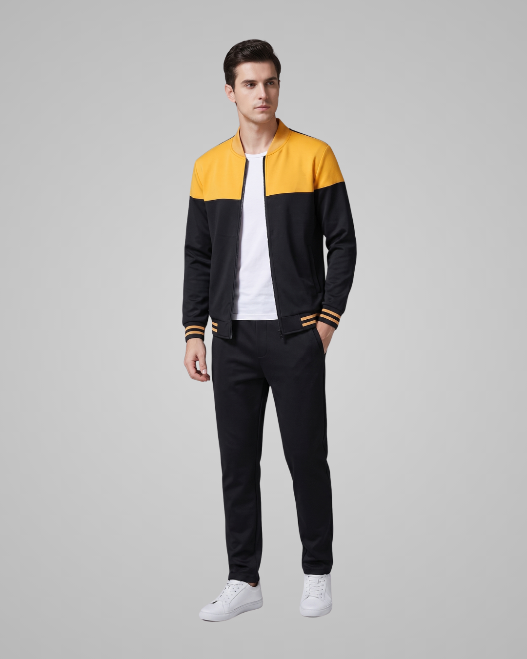 A man modeling a color-blocked track jacket with a yellow yoke and black sleeves, paired with black pants and white sneakers, against a solid gray studio background.