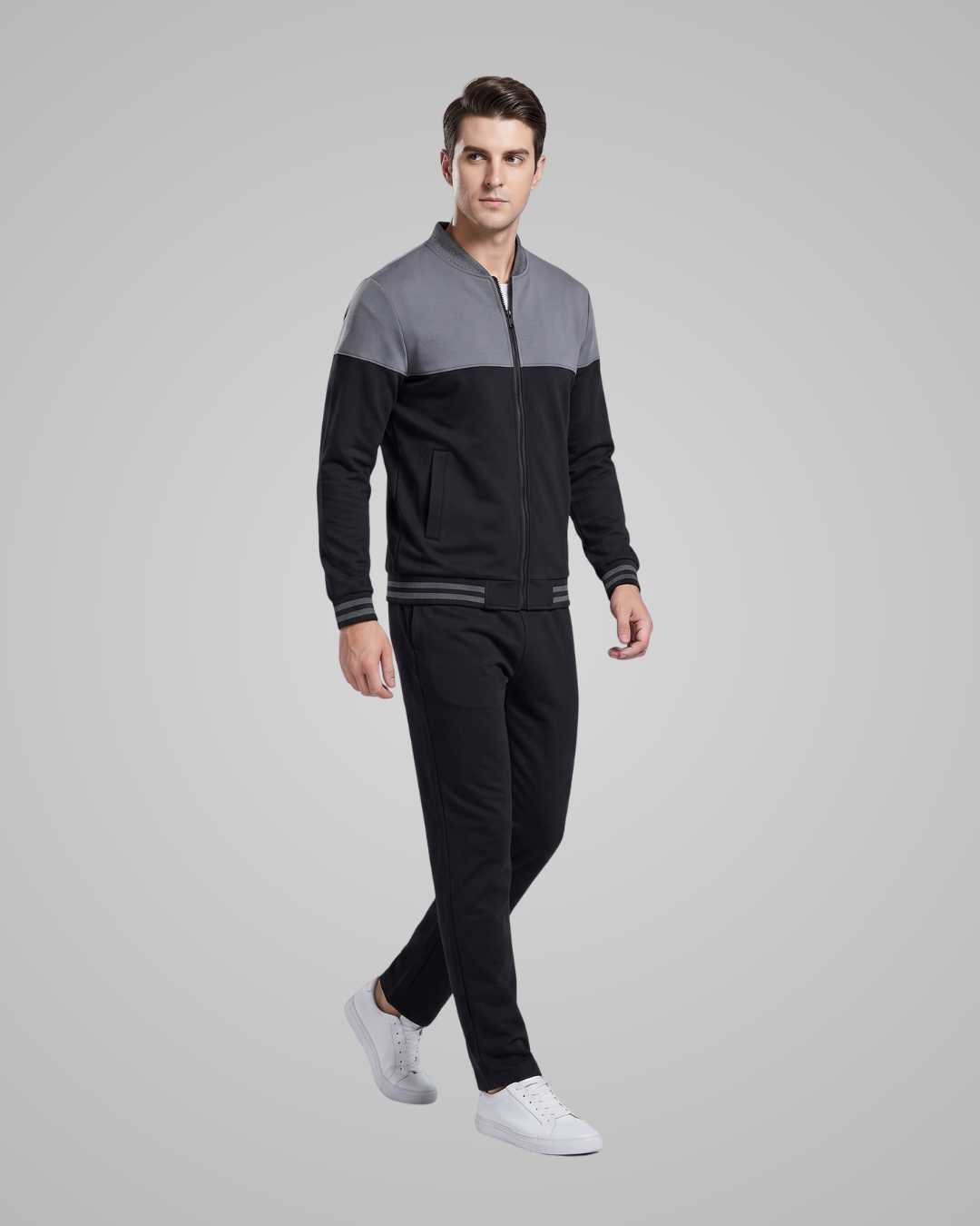 A man modeling a two-tone colorblock zip-up jacket and matching black track pants against a solid gray studio background.