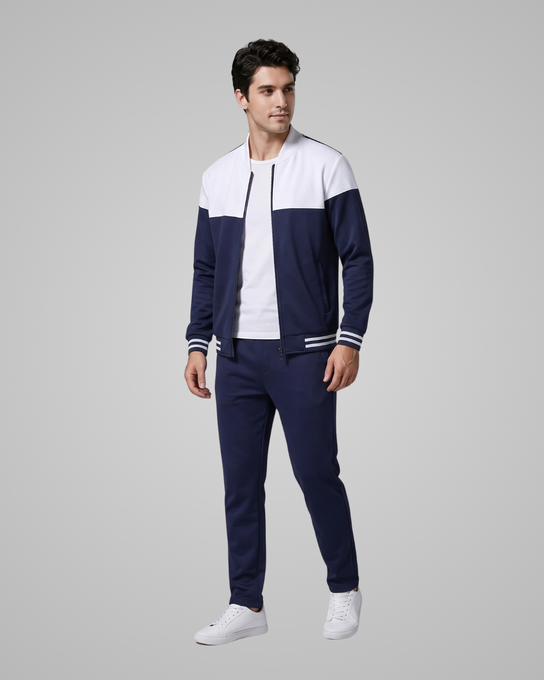 A man modeling a navy blue and white color-blocked track jacket and matching navy pants, styled with a white t-shirt and white sneakers, against a solid gray studio background.