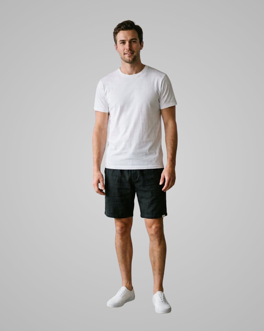 A man modeling a plain white short-sleeve crewneck t-shirt, paired with black shorts and white sneakers, standing against a solid light gray background.
