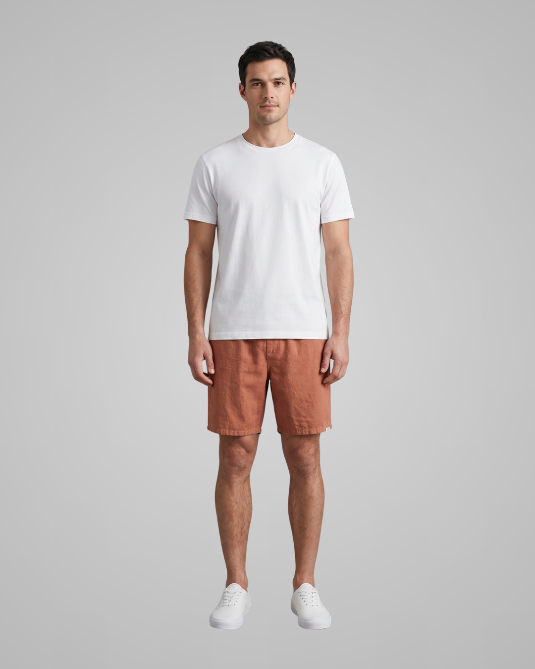 A man modeling a white crew-neck t-shirt and rust-orange shorts against a solid gray studio background.