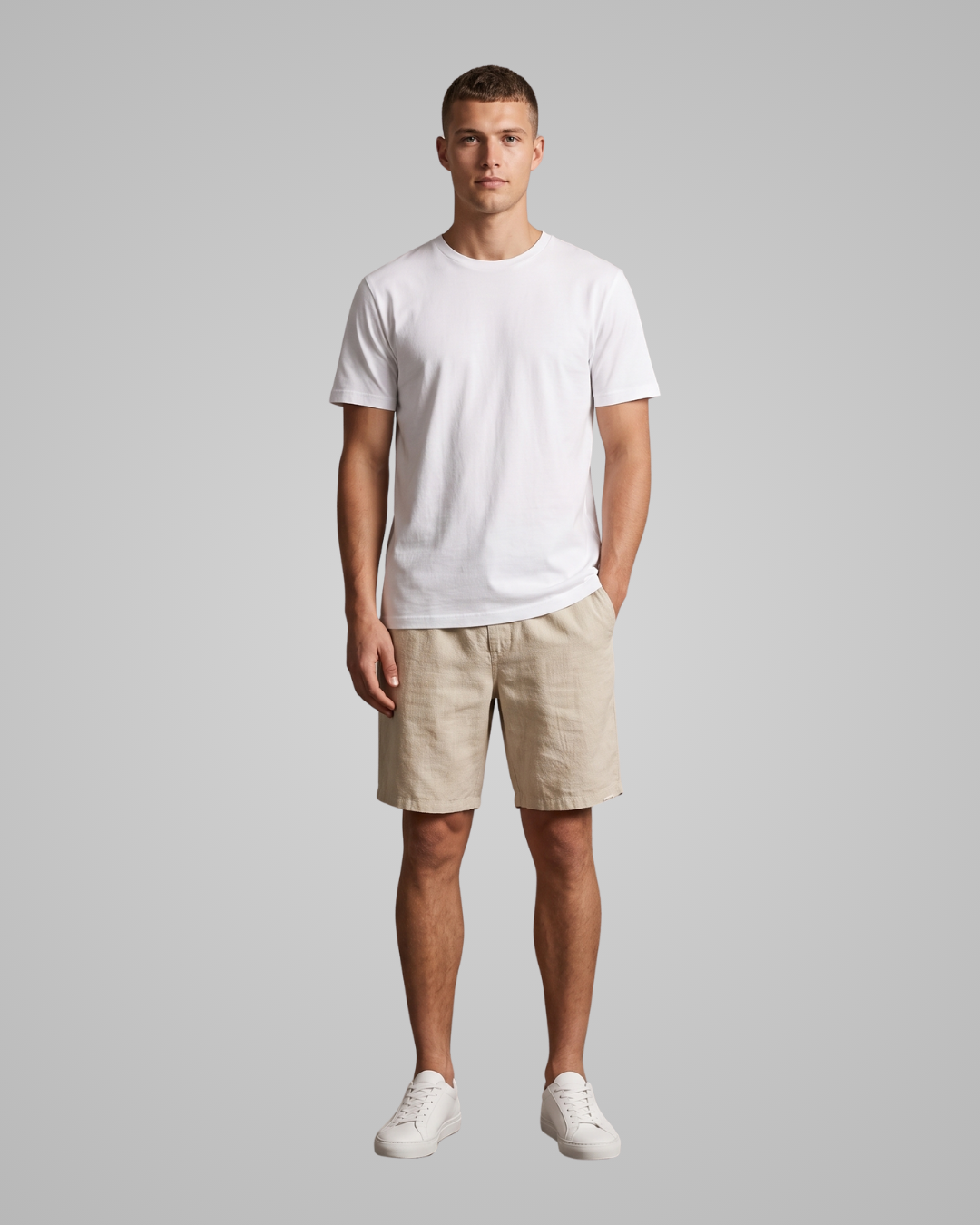 A man modeling a white short-sleeve crewneck t-shirt, beige linen shorts, and white sneakers against a solid gray studio background.
