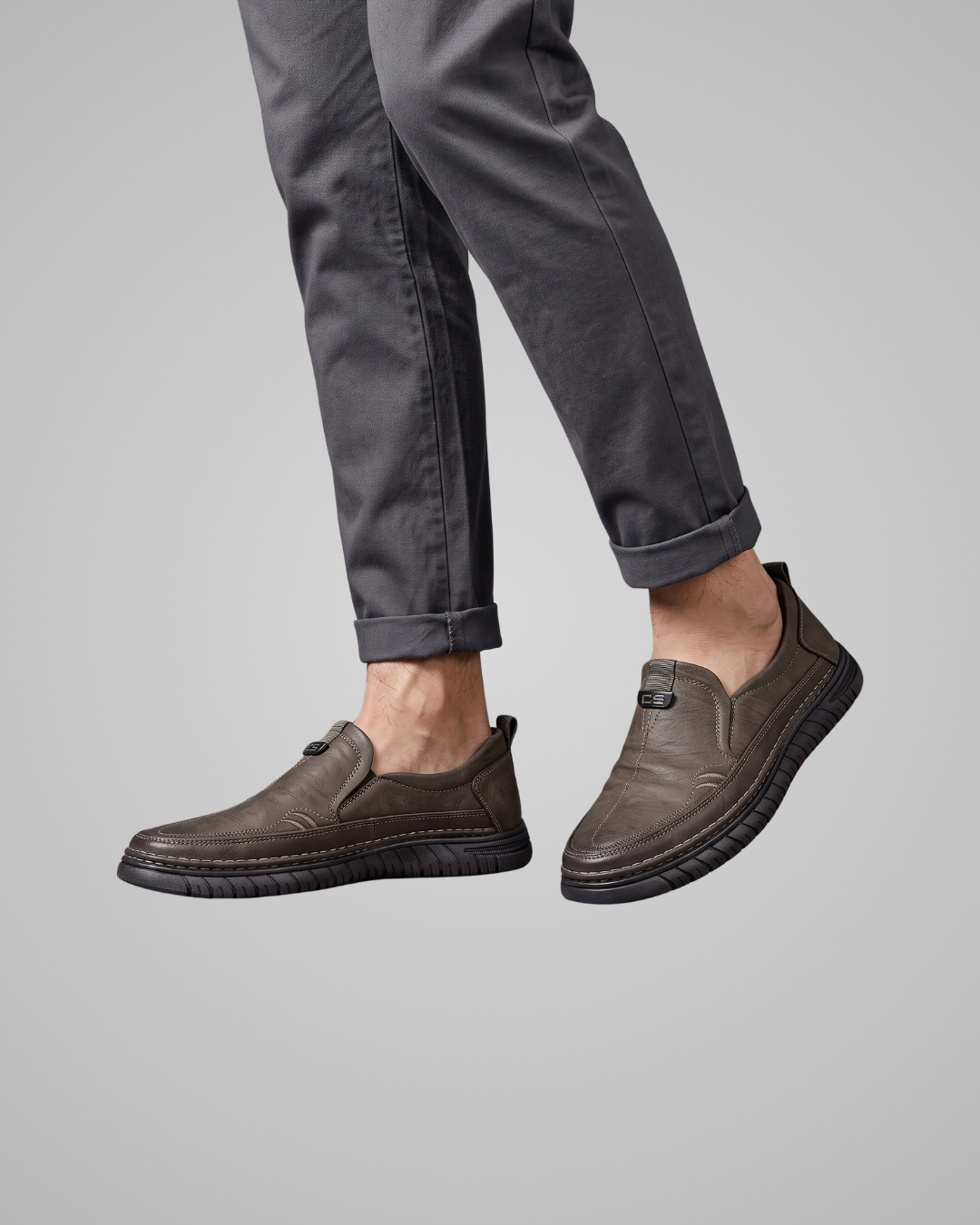 A person wearing dark gray cuffed pants and dark brown slip-on loafers with textured soles, shown against a solid gray background.