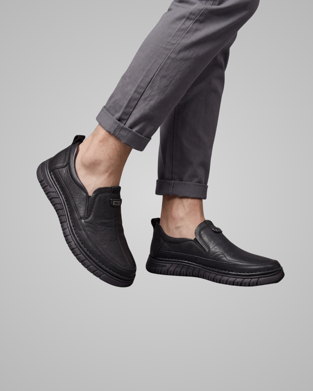 A pair of black slip-on leather shoes worn with dark gray cuffed trousers, shown against a solid light gray background.