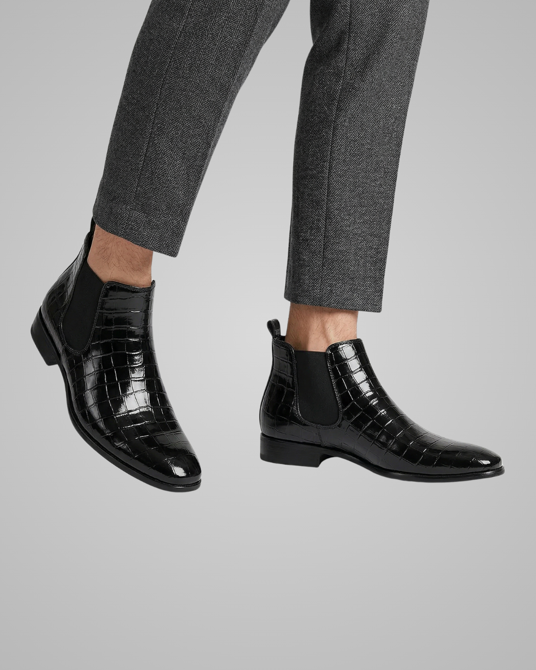 Black crocodile-patterned Chelsea boots worn with gray pants on a light gray background