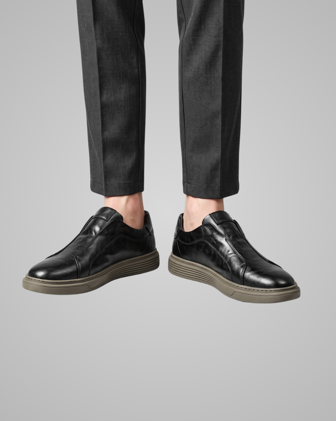 A pair of men's black leather lace-up shoes with textured soles, shown on a model wearing dark charcoal trousers against a solid gray background.