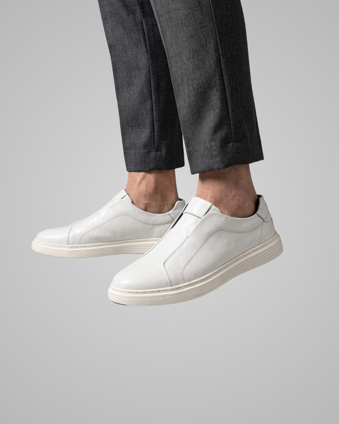 White leather slip-on sneakers worn with dark gray trousers, shown from the feet up against a solid gray background.