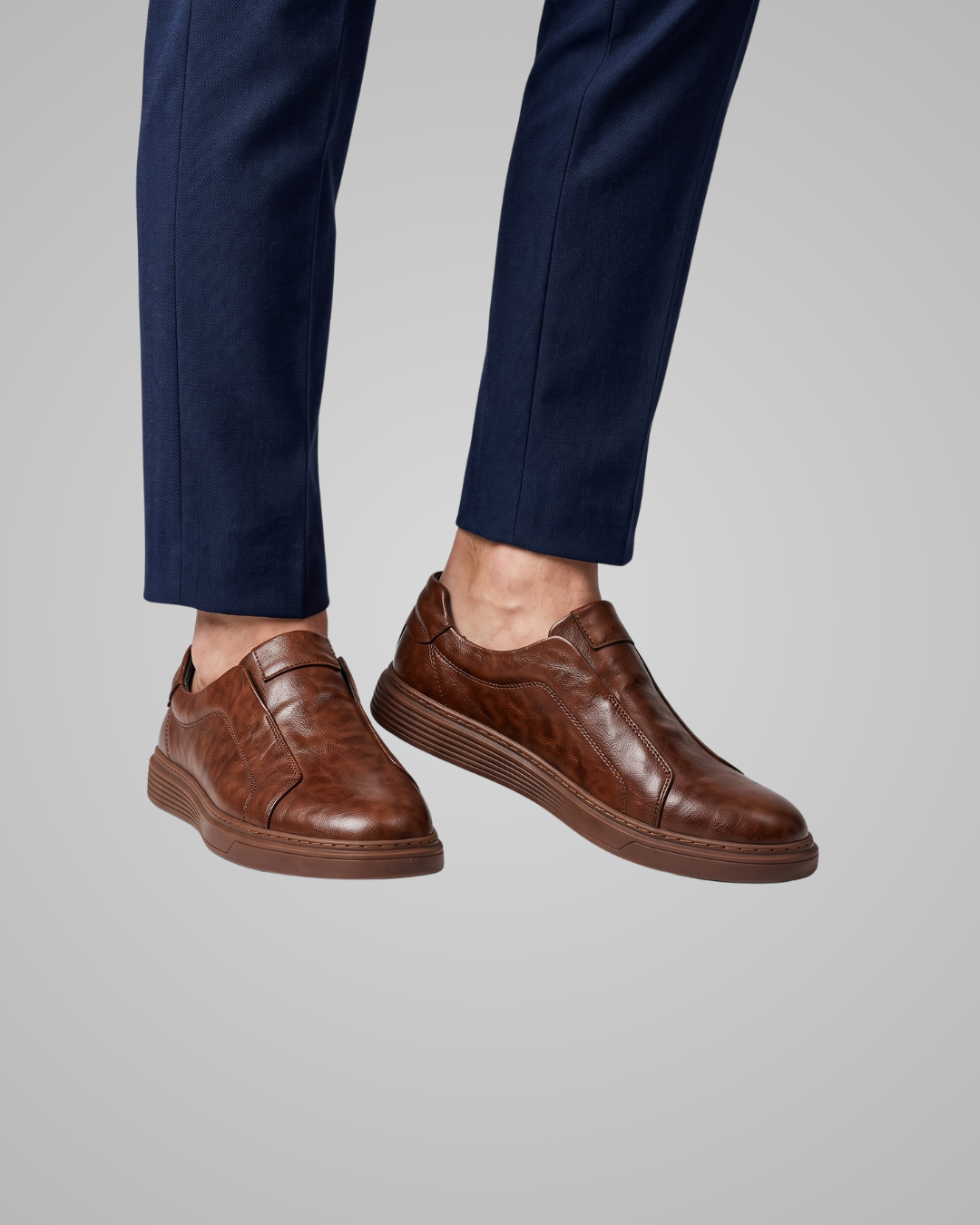 A pair of men's brown leather slip-on shoes with a textured finish and low stacked heels, shown on a model wearing navy blue trousers against a solid gray background.
