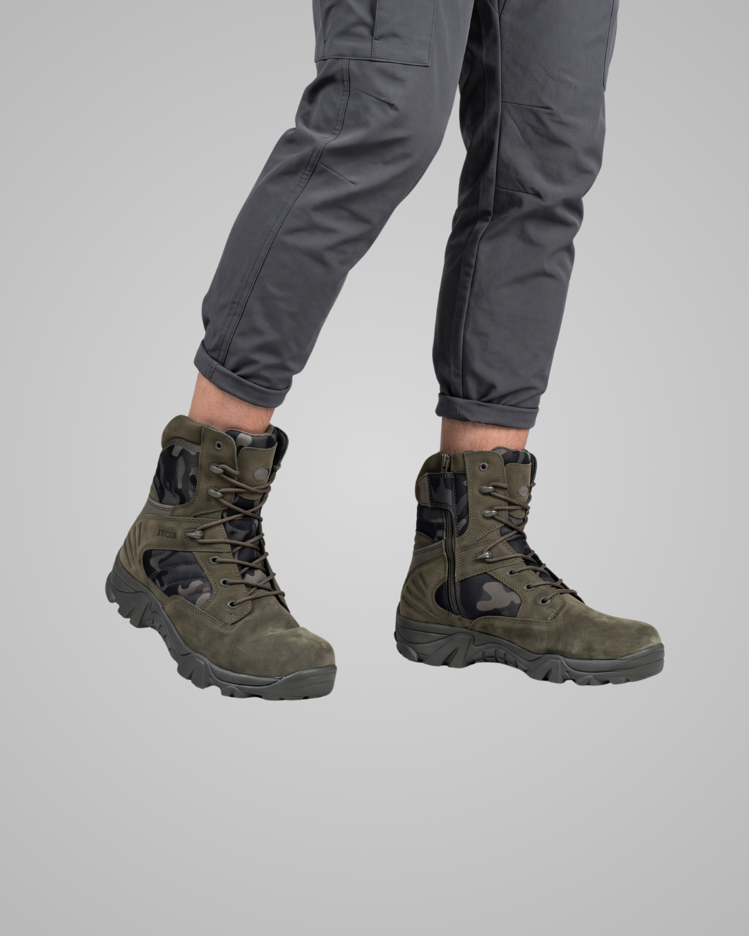 A pair of olive green tactical combat boots with black accents, shown on a model wearing dark gray pants against a solid gray background.