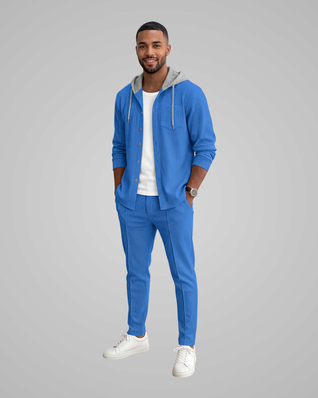 A man modeling a bright blue two-piece lounge set consisting of a zip-up hoodie with a gray hood lining and matching blue pants, worn over a white t-shirt and white sneakers, against a solid gray background.