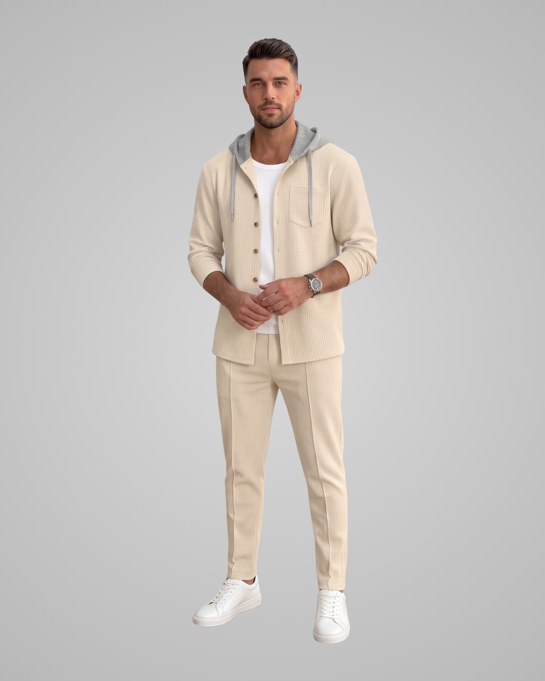 A man modeling a light beige two-piece lounge set consisting of a hooded button-front cardigan with a gray hood lining and matching slim-fit trousers, worn over a white t-shirt and white sneakers, against a solid gray background.