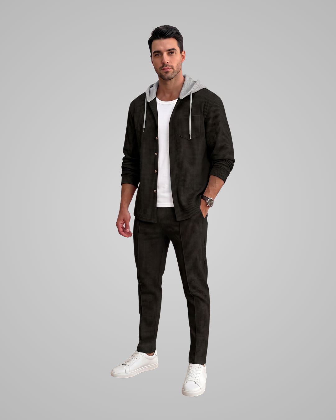 A man modeling a black zip-up hoodie with a gray hood lining, worn open over a white t-shirt and black trousers, paired with white sneakers, against a solid gray background.