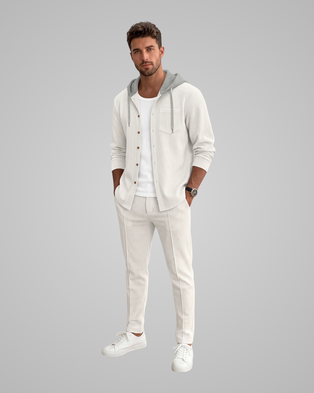 A man modeling a light cream-colored loungewear set consisting of a button-up hoodie with a gray hood, matching jogger pants, and white sneakers, set against a solid gray background.
