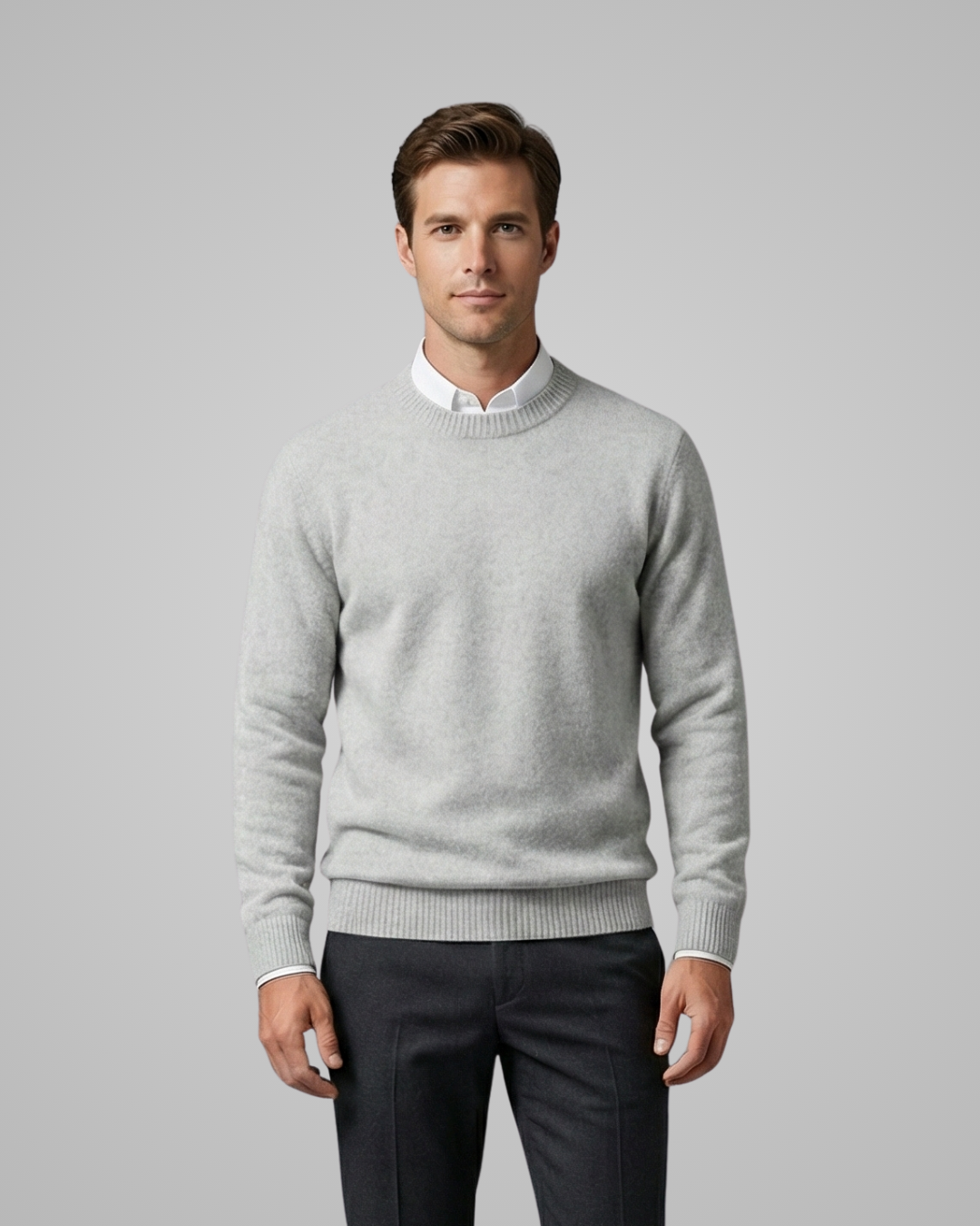 Man wearing a light gray sweater over a white shirt with dark pants on a plain background