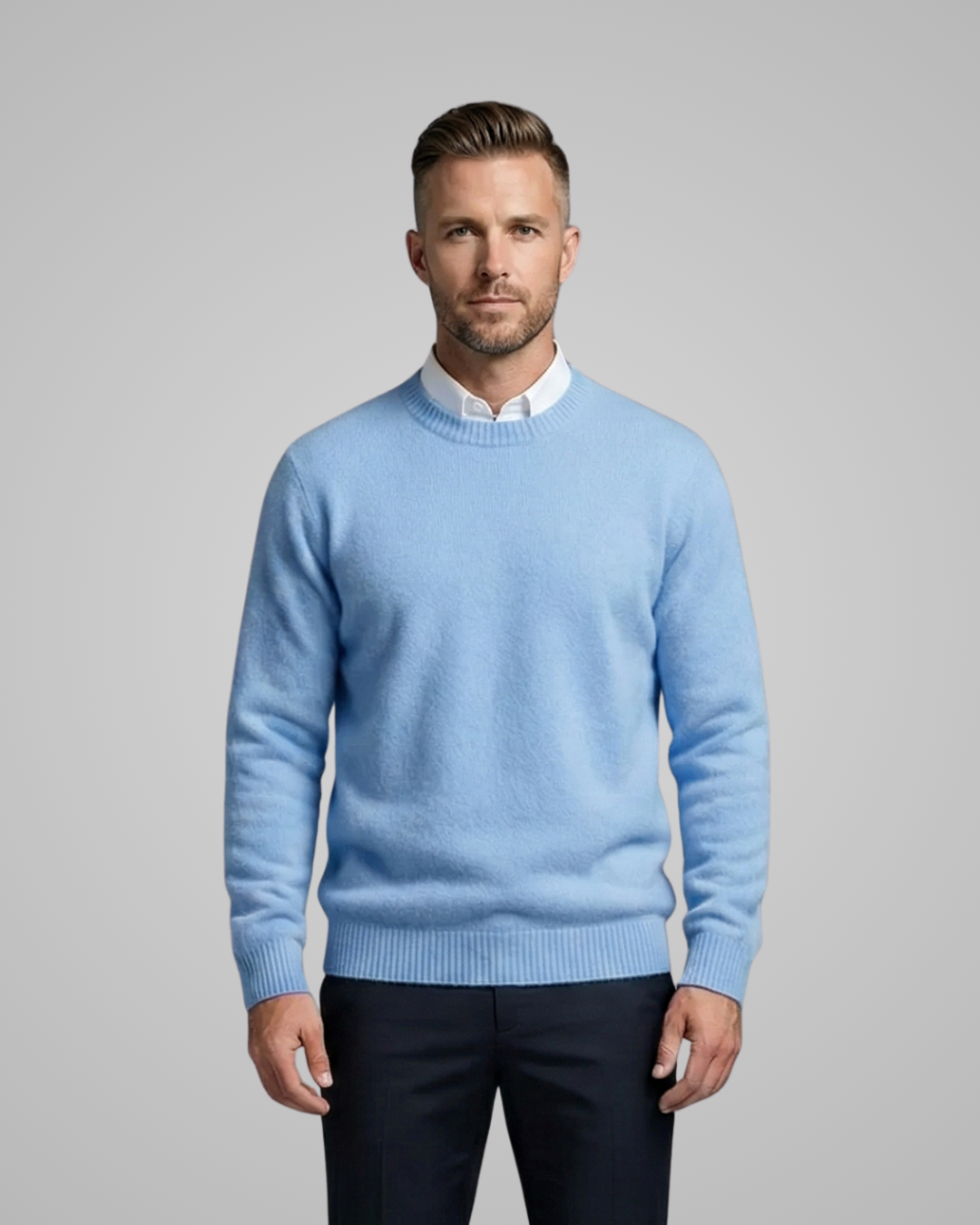 Man wearing a light blue sweater over a white shirt with dark pants on a gray background