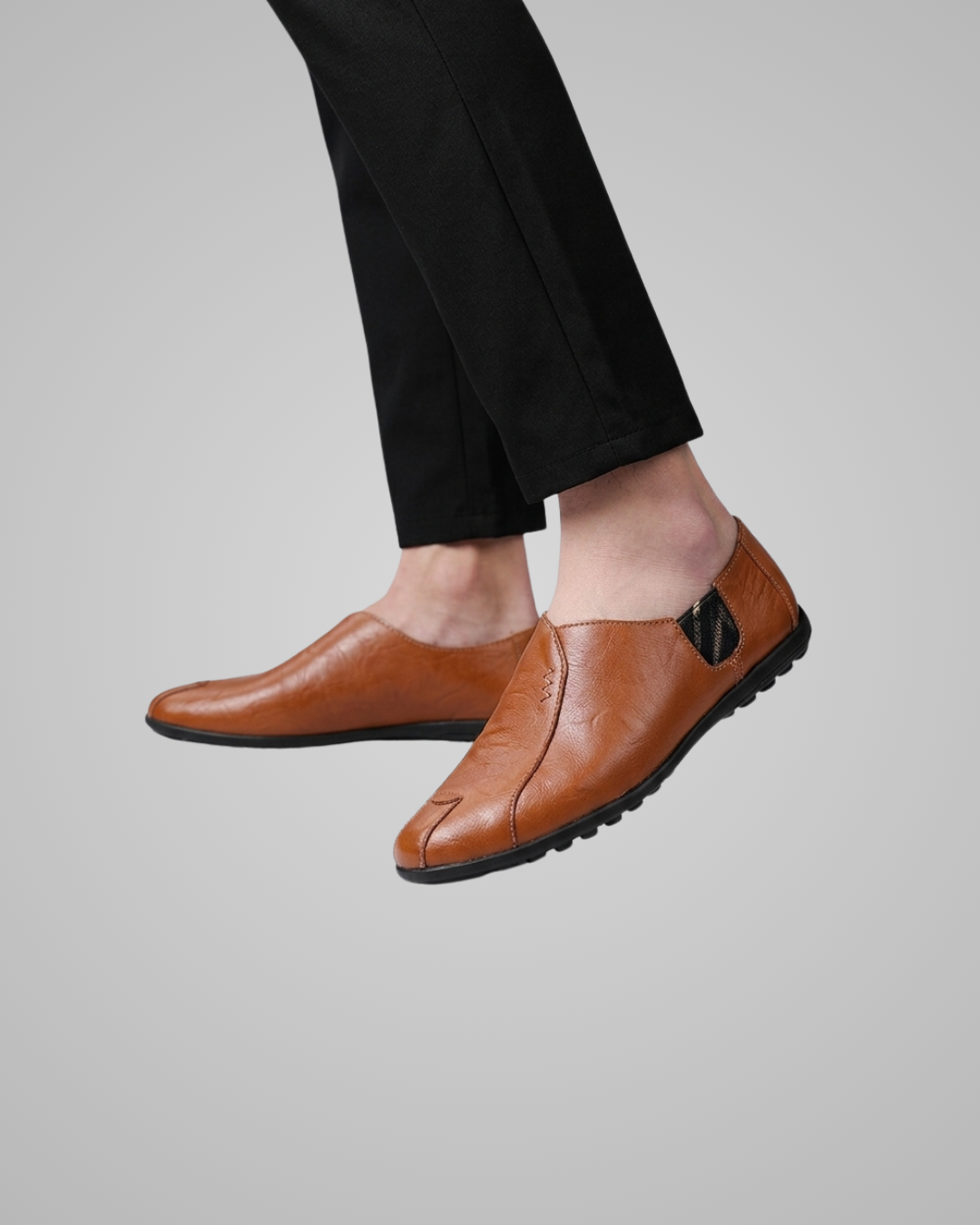 A pair of men's brown leather slip-on loafers with black soles, shown on a model's feet wearing black trousers against a solid gray background.