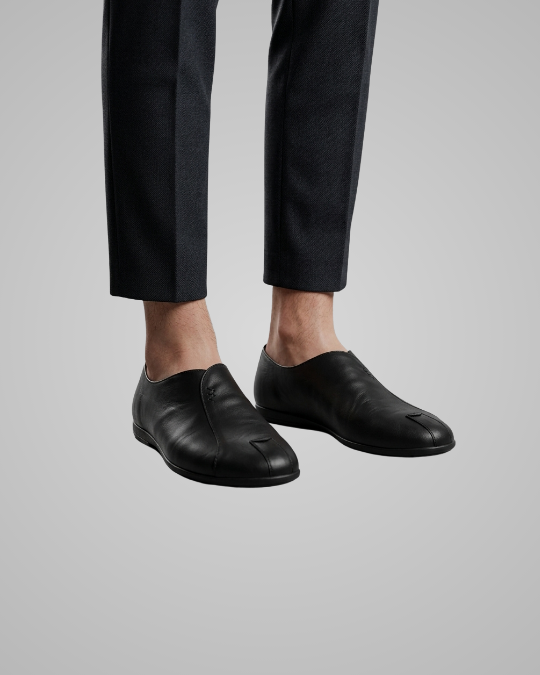 A close-up of a person's feet wearing black leather slip-on loafers with a distinctive cutout detail on the toe, paired with black trousers, against a solid gray background.