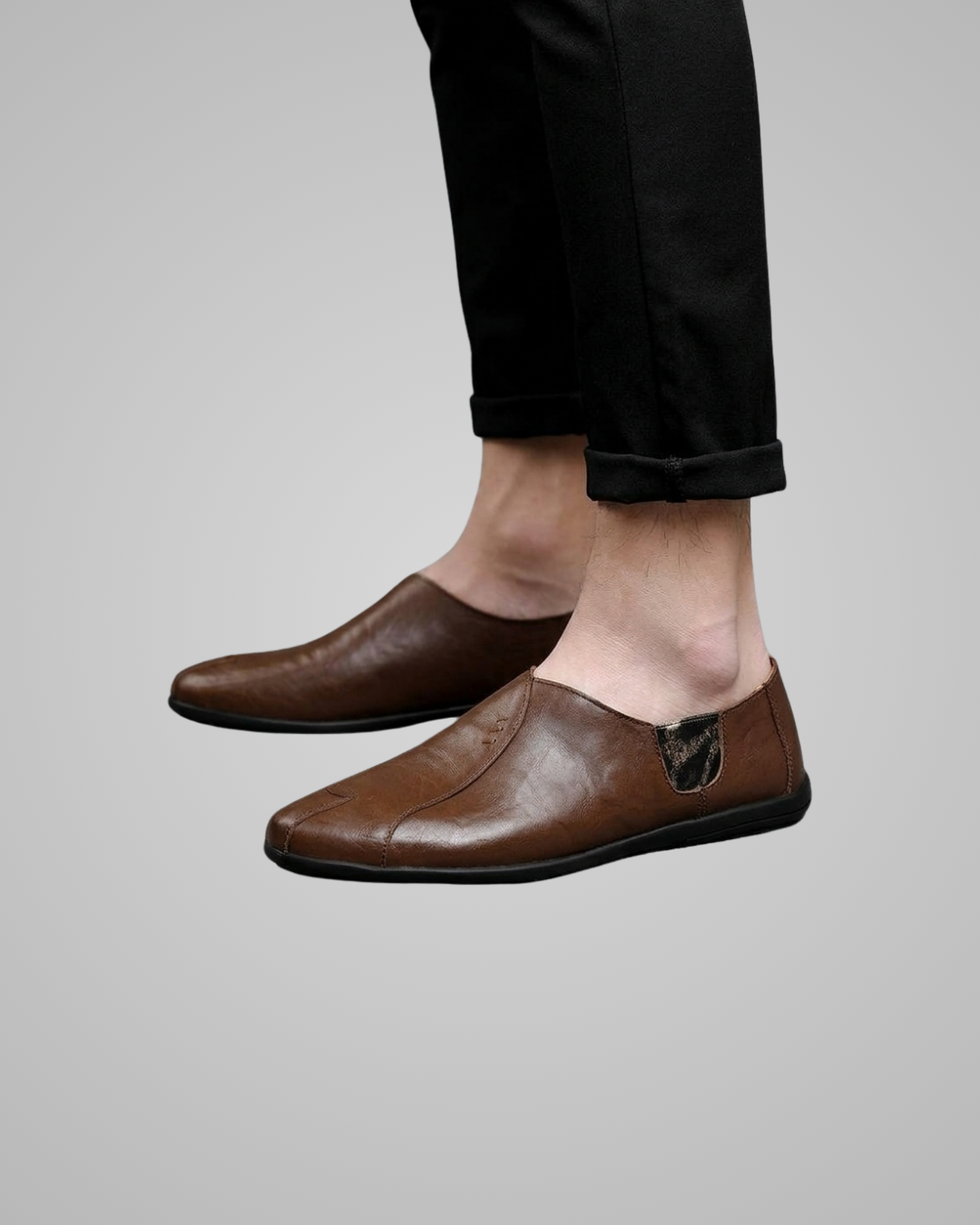 A pair of men's brown leather slip-on loafers shown on a model's feet, paired with black cuffed trousers against a solid gray background.