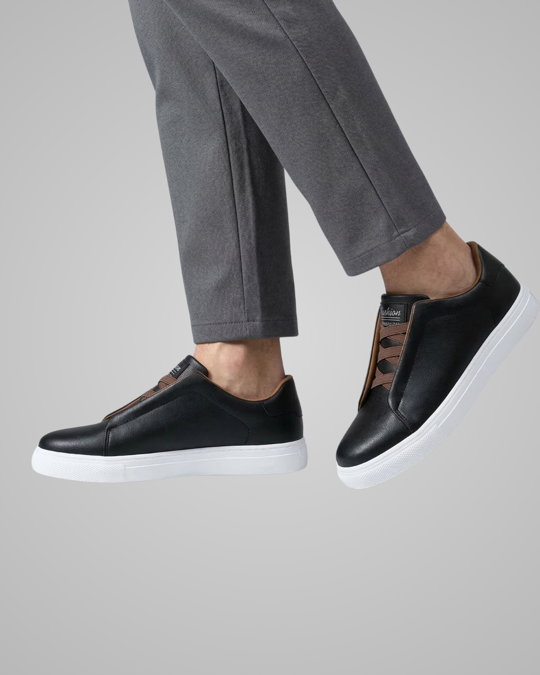 Black slip-on sneakers with white soles worn with gray pants on a light gray background