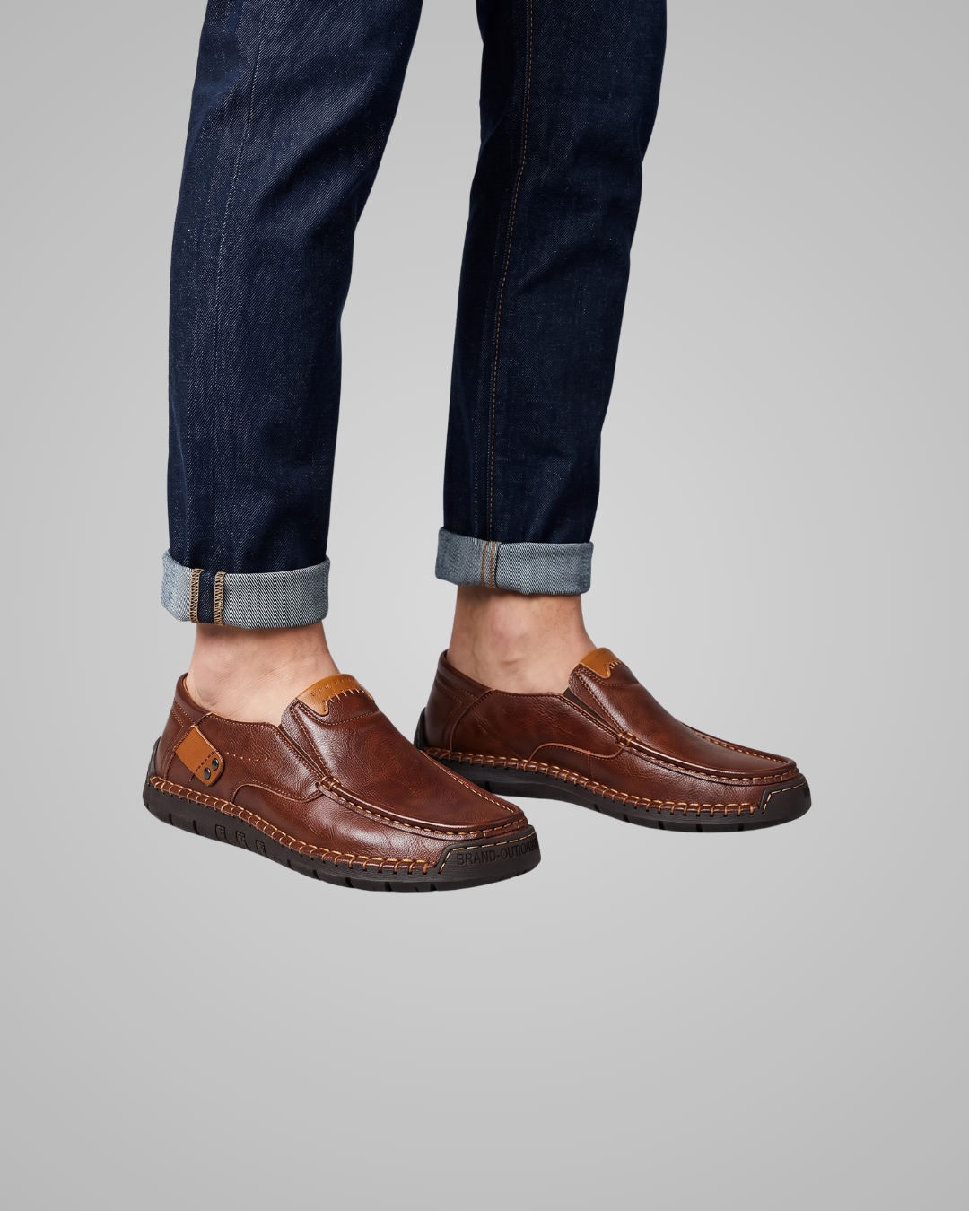 A pair of men's brown leather slip-on loafers with contrast stitching and a small tan leather accent tab, shown on a model wearing dark blue jeans against a solid gray background.