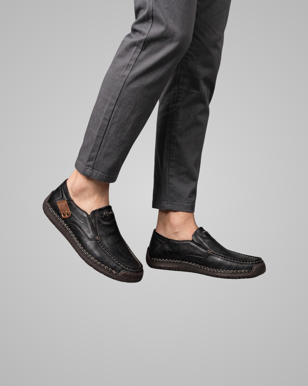 A person wearing black leather slip-on loafers with dark gray trousers, shown from the calves down against a solid gray background.
