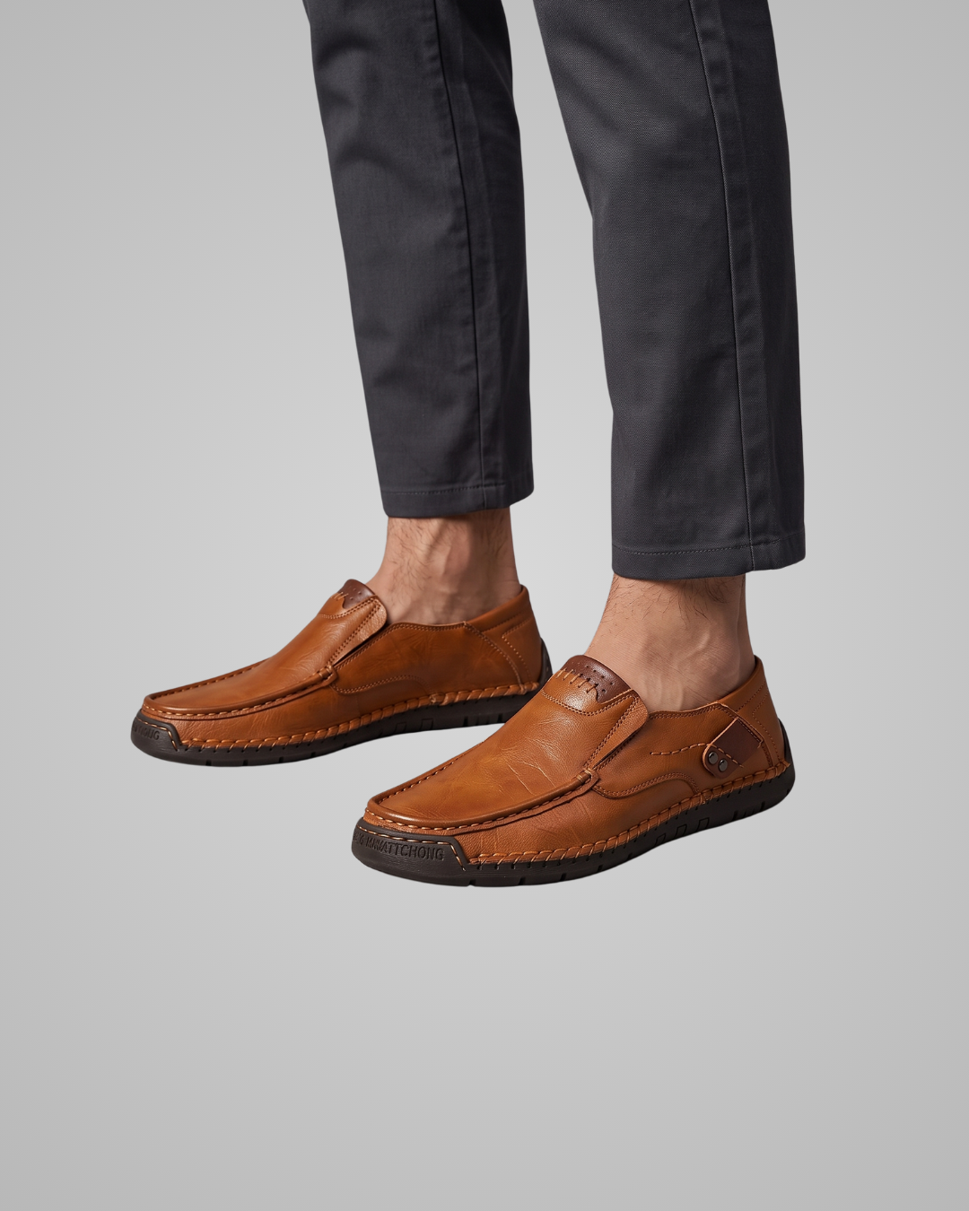 A person wearing dark charcoal pants and tan leather slip-on loafers with dark rubber soles, shown from the calves down against a plain light gray studio background.