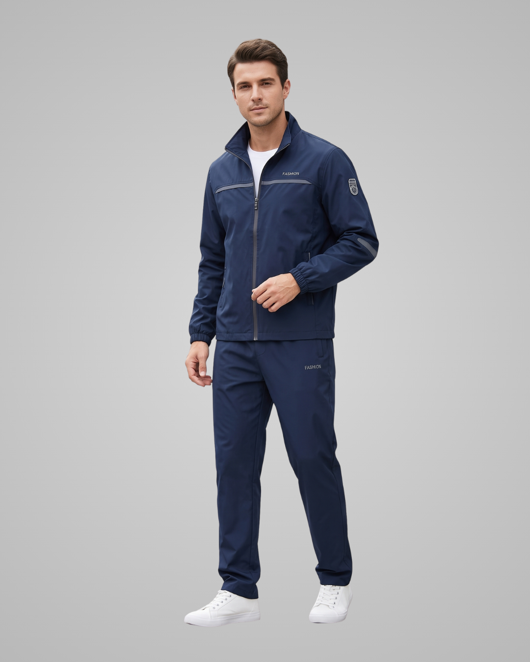 A man modeling a navy blue athletic tracksuit, consisting of a zip-up jacket and matching pants, paired with white sneakers, against a solid gray studio background.