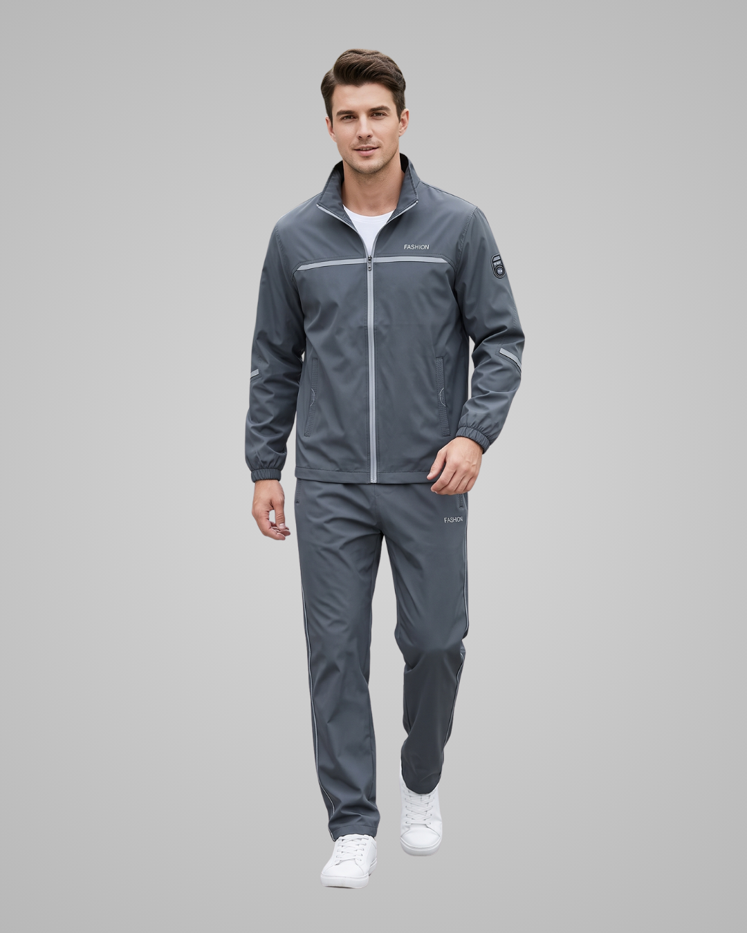 A man modeling a dark gray athletic tracksuit, consisting of a zip-up jacket and matching pants, with white sneakers, against a solid gray studio background.