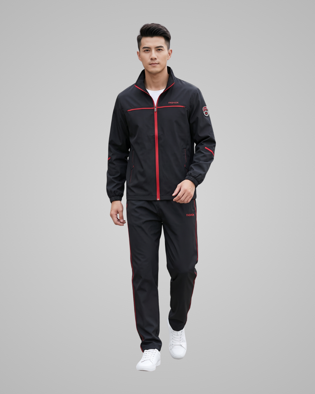 A man modeling a black athletic tracksuit with red zipper and piping details, consisting of a zip-up jacket and matching pants, paired with white sneakers against a solid gray background.