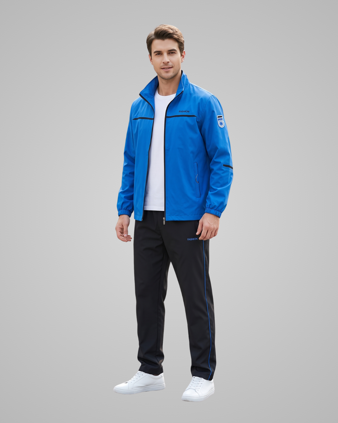 A man modeling a bright blue athletic jacket over a white t-shirt, paired with black track pants and white sneakers, against a solid gray studio background.