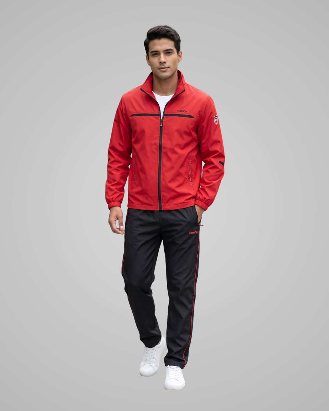 A man modeling a red zip-up athletic jacket with black trim and black track pants with red side stripes, paired with white sneakers, against a solid gray background.
