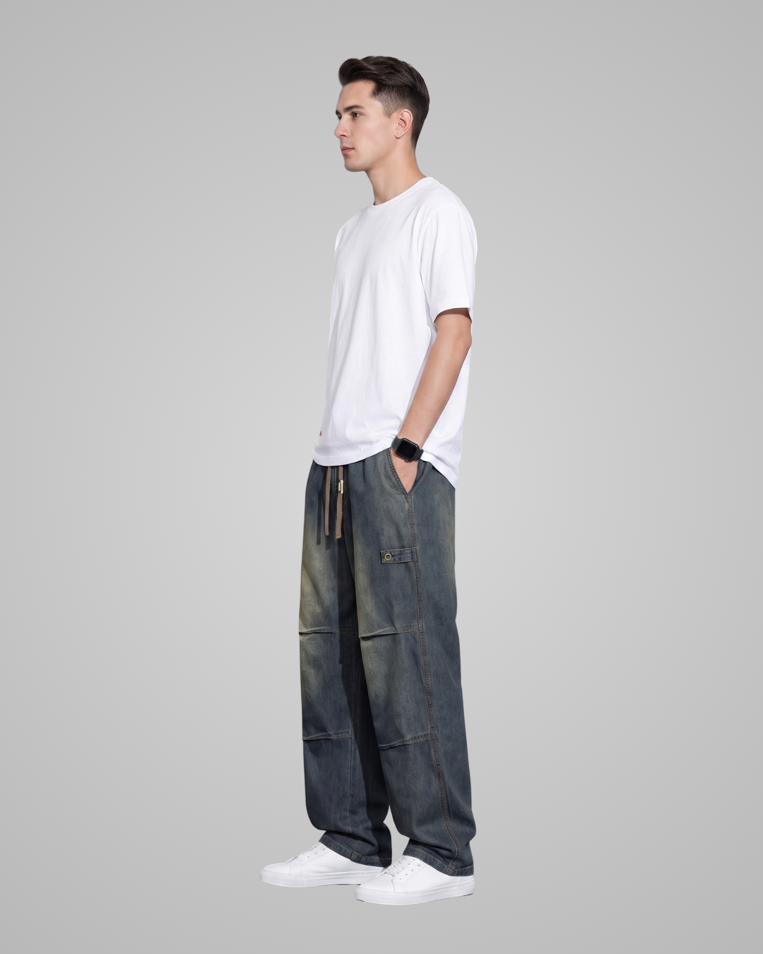 A man in profile wearing a white t-shirt, dark gray baggy cargo pants, and white sneakers, standing against a solid gray background.