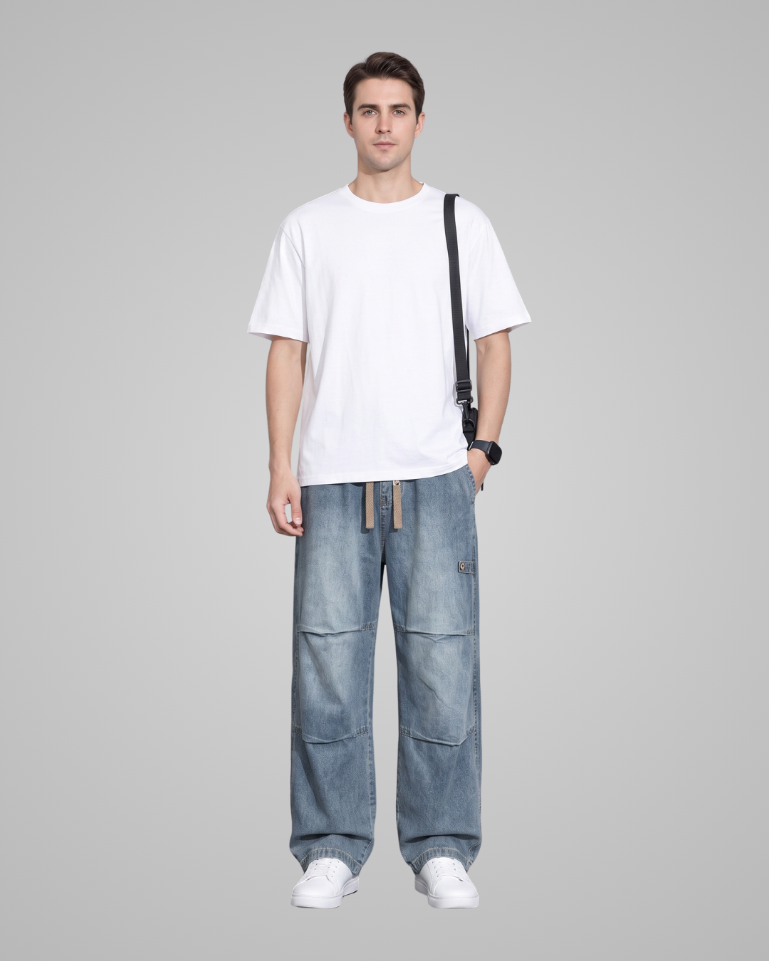 A man modeling a white crew-neck t-shirt, wide-leg light blue denim cargo pants, and white sneakers, with a black shoulder bag strap visible against a solid gray studio background.