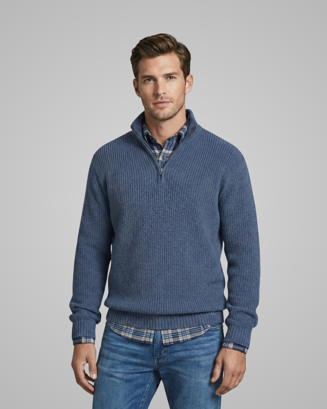Man wearing a blue sweater over a plaid shirt with jeans on a gray background