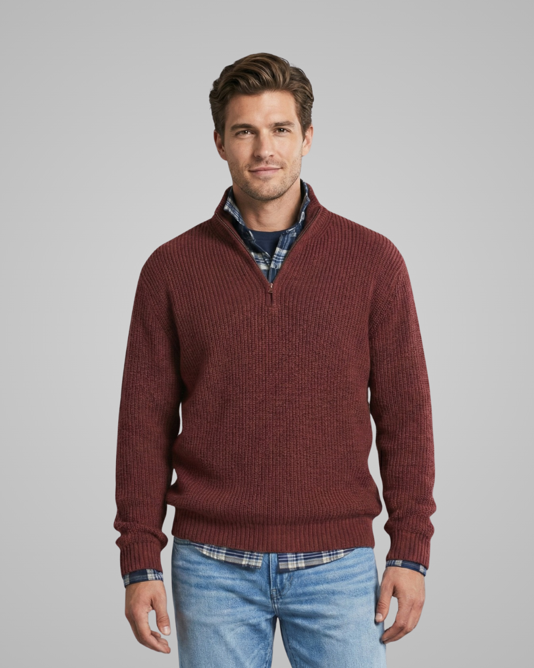 Man wearing a burgundy sweater over a plaid shirt with light blue jeans on a gray background