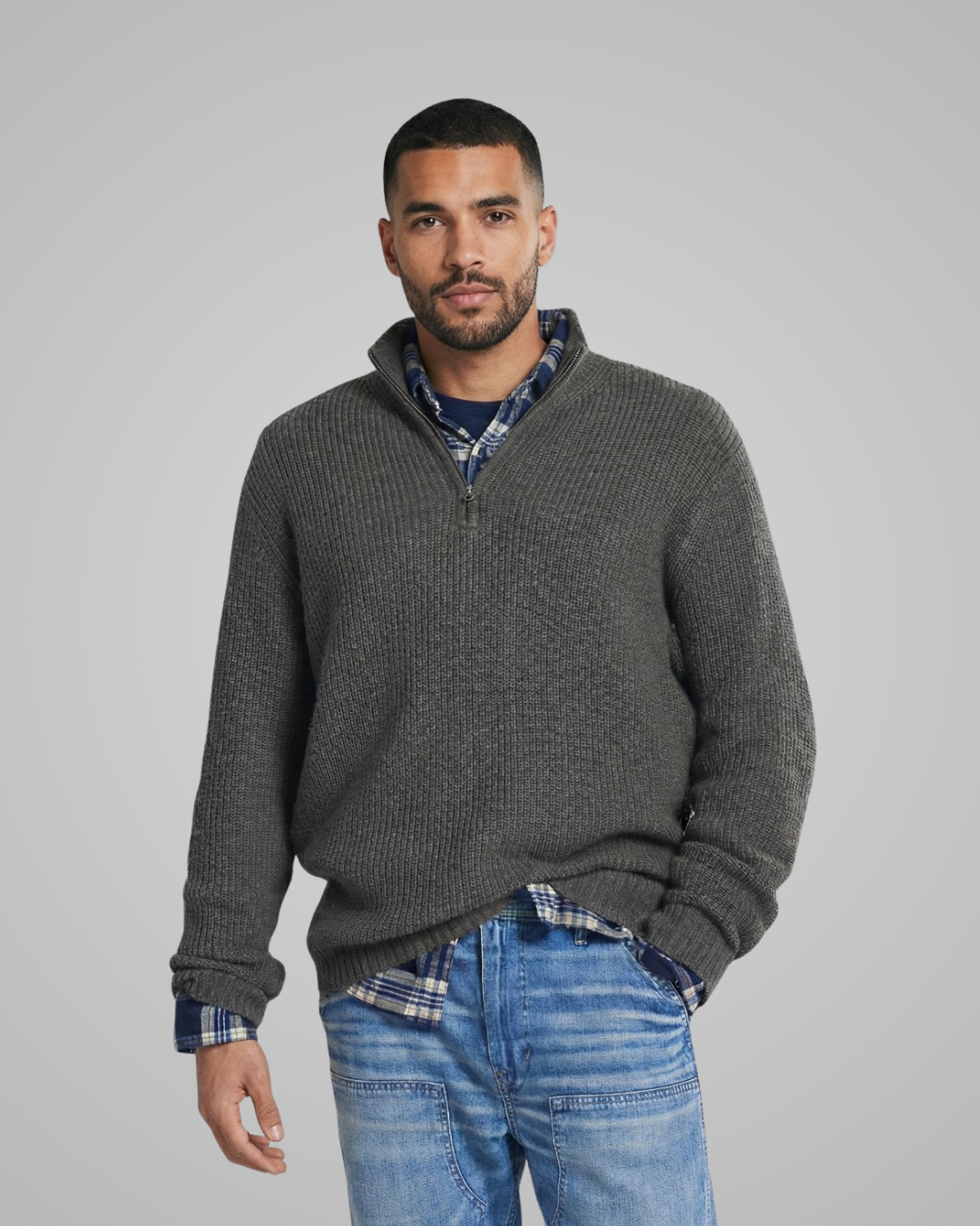 Man wearing a gray sweater over a plaid shirt with blue jeans on a gray background