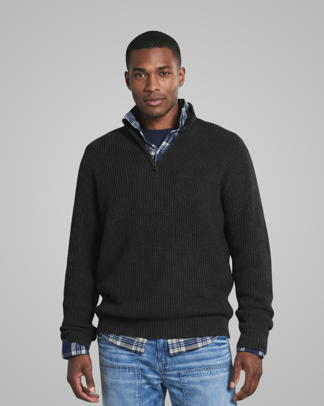 Man wearing a black sweater over a plaid shirt with blue jeans on a gray background