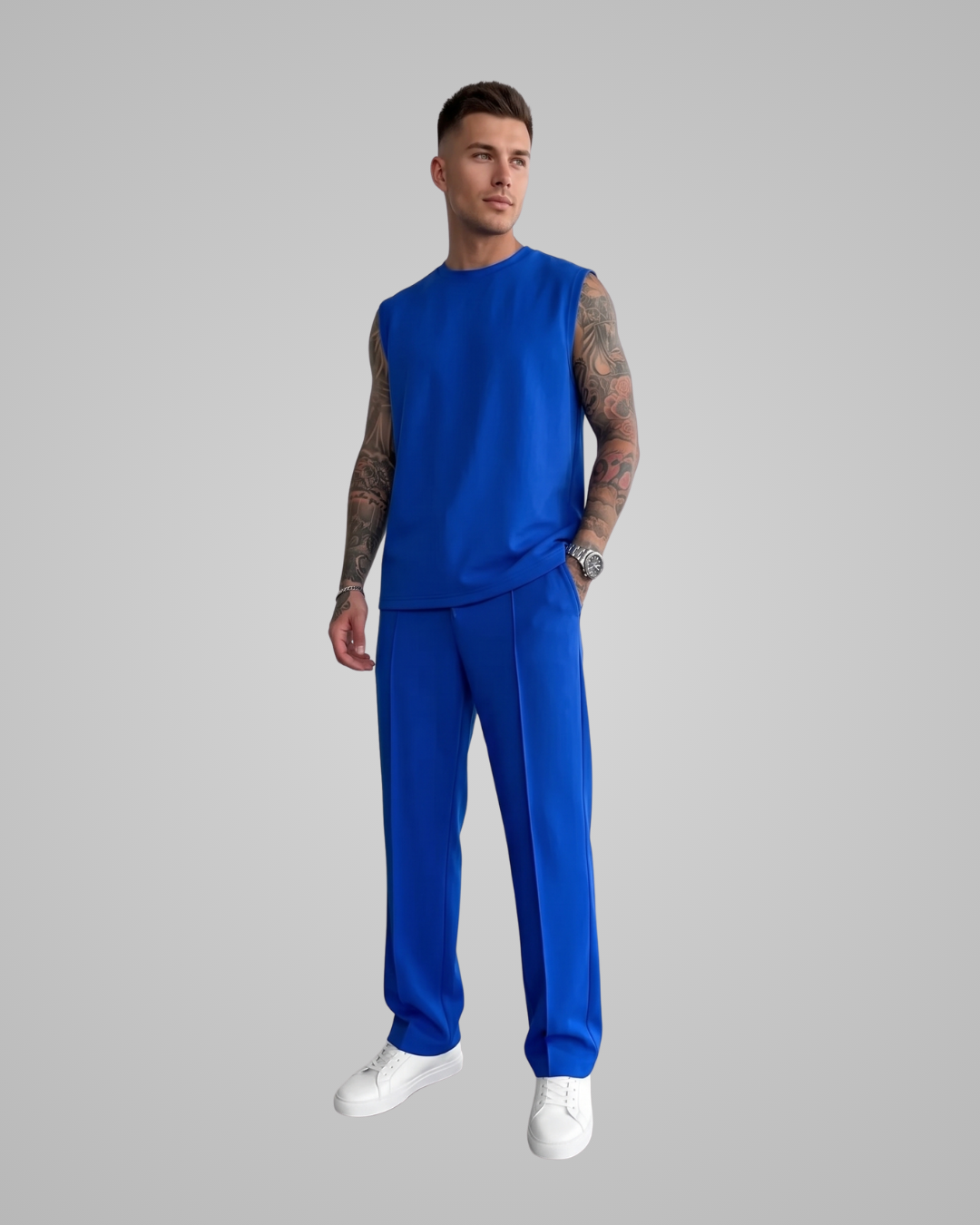 A man modeling a matching royal blue lounge set consisting of a sleeveless crewneck top and wide-leg trousers, paired with white sneakers, against a solid gray studio background.