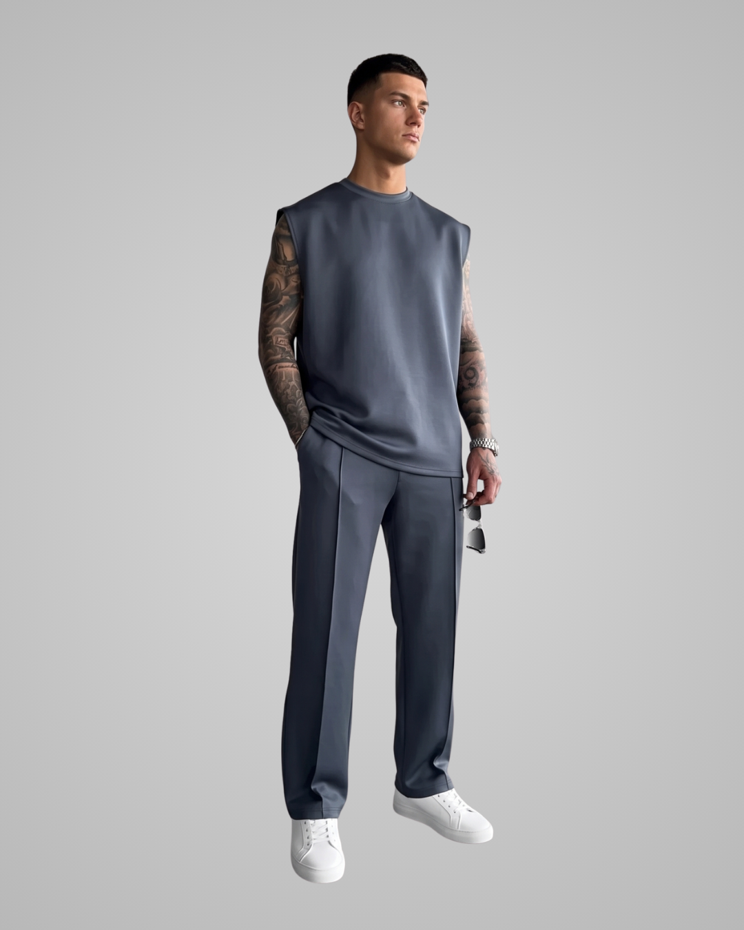 A man modeling a sleeveless dark slate blue top and matching trousers with white sneakers, set against a solid light gray background.