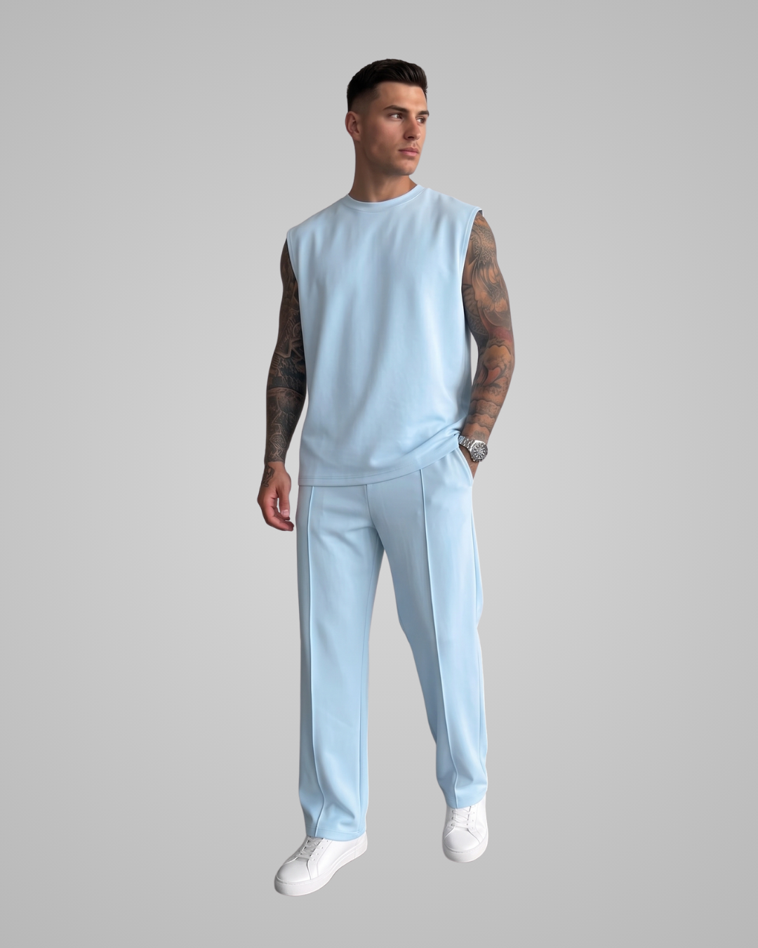 A man modeling a light blue sleeveless crewneck top and matching light blue trousers, paired with white sneakers, against a solid gray background.
