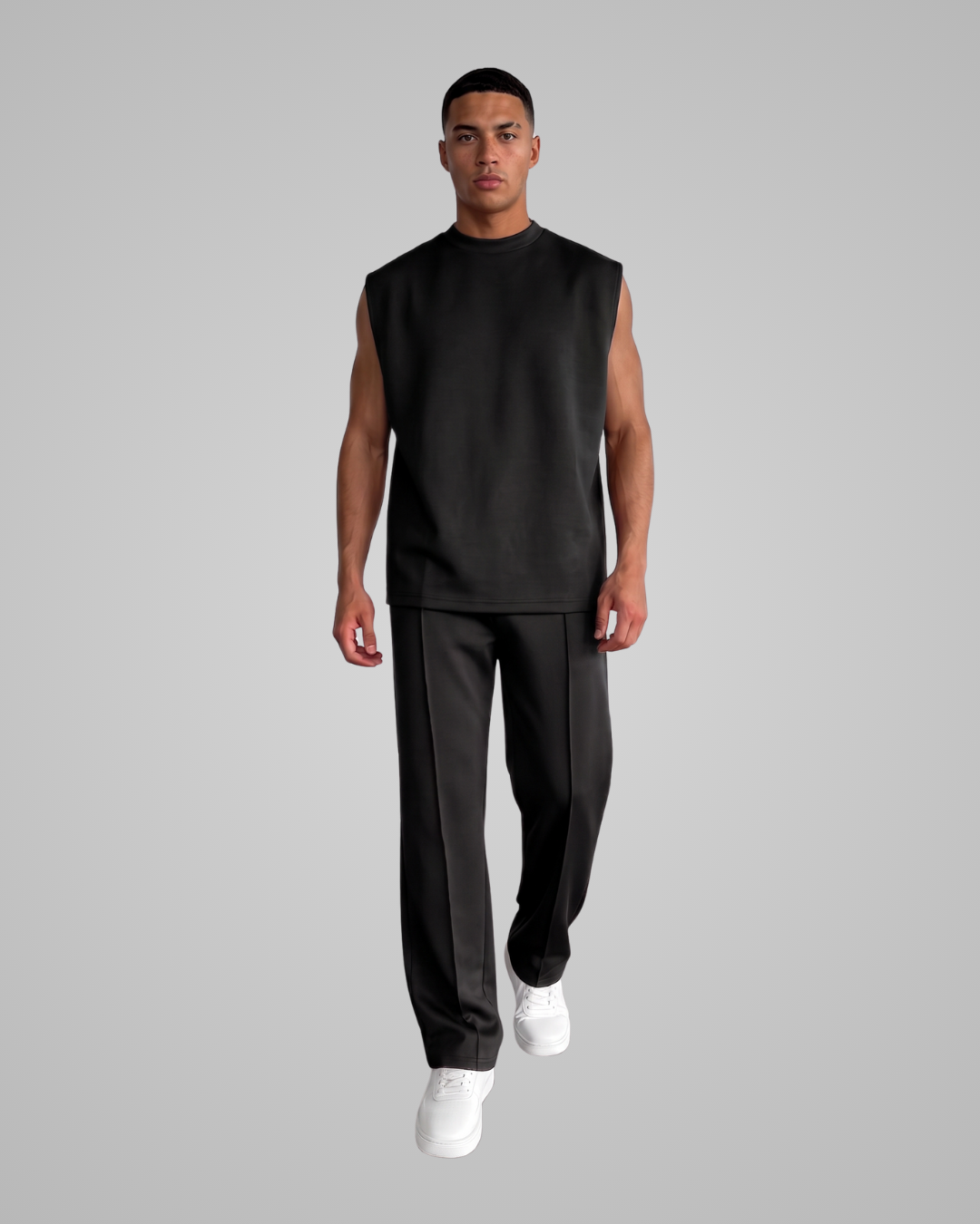 A man modeling a black sleeveless mock-neck top and black pants with white sneakers, set against a solid gray studio background.