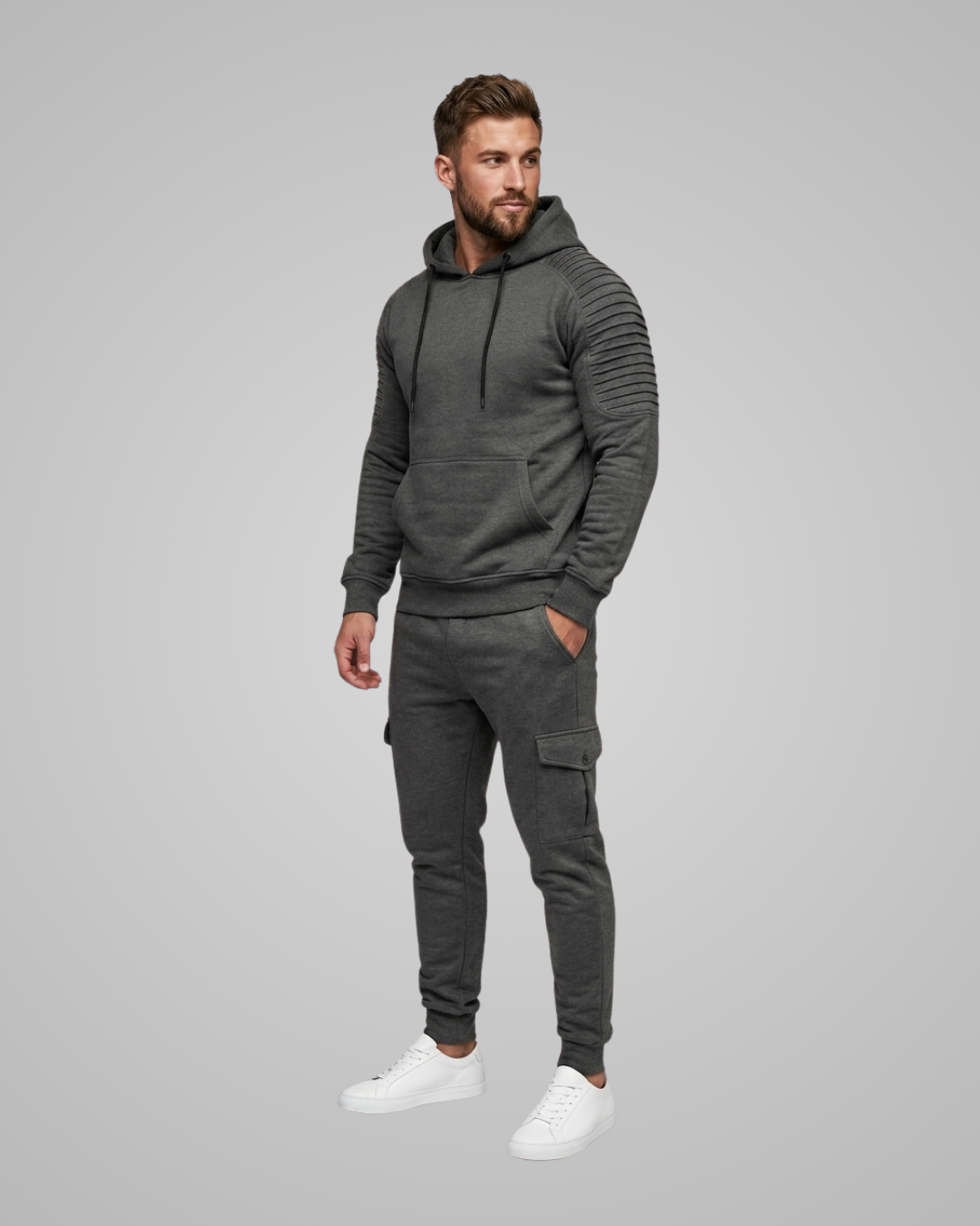 A man modeling a dark gray ribbed hoodie and matching cargo jogger pants with white sneakers, standing against a solid gray studio background.