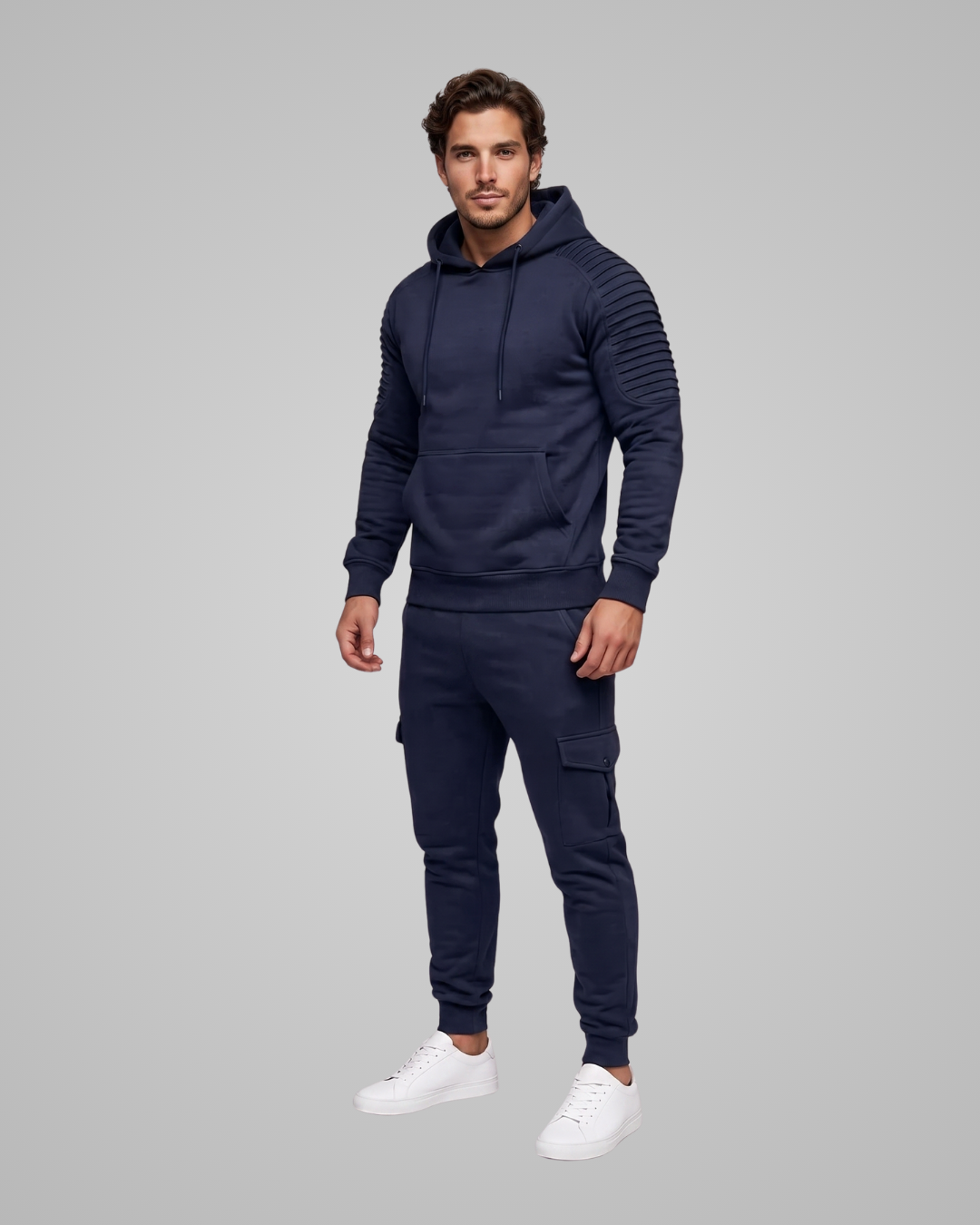 A man modeling a matching navy blue hoodie and jogger pants set with white sneakers, standing against a solid gray studio background.