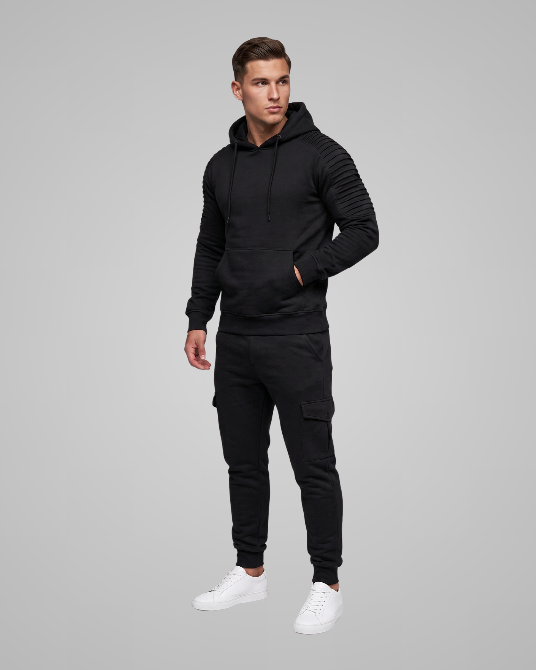 A man modeling a black ribbed knit hoodie and matching black cargo jogger pants with white sneakers, set against a solid gray studio background.