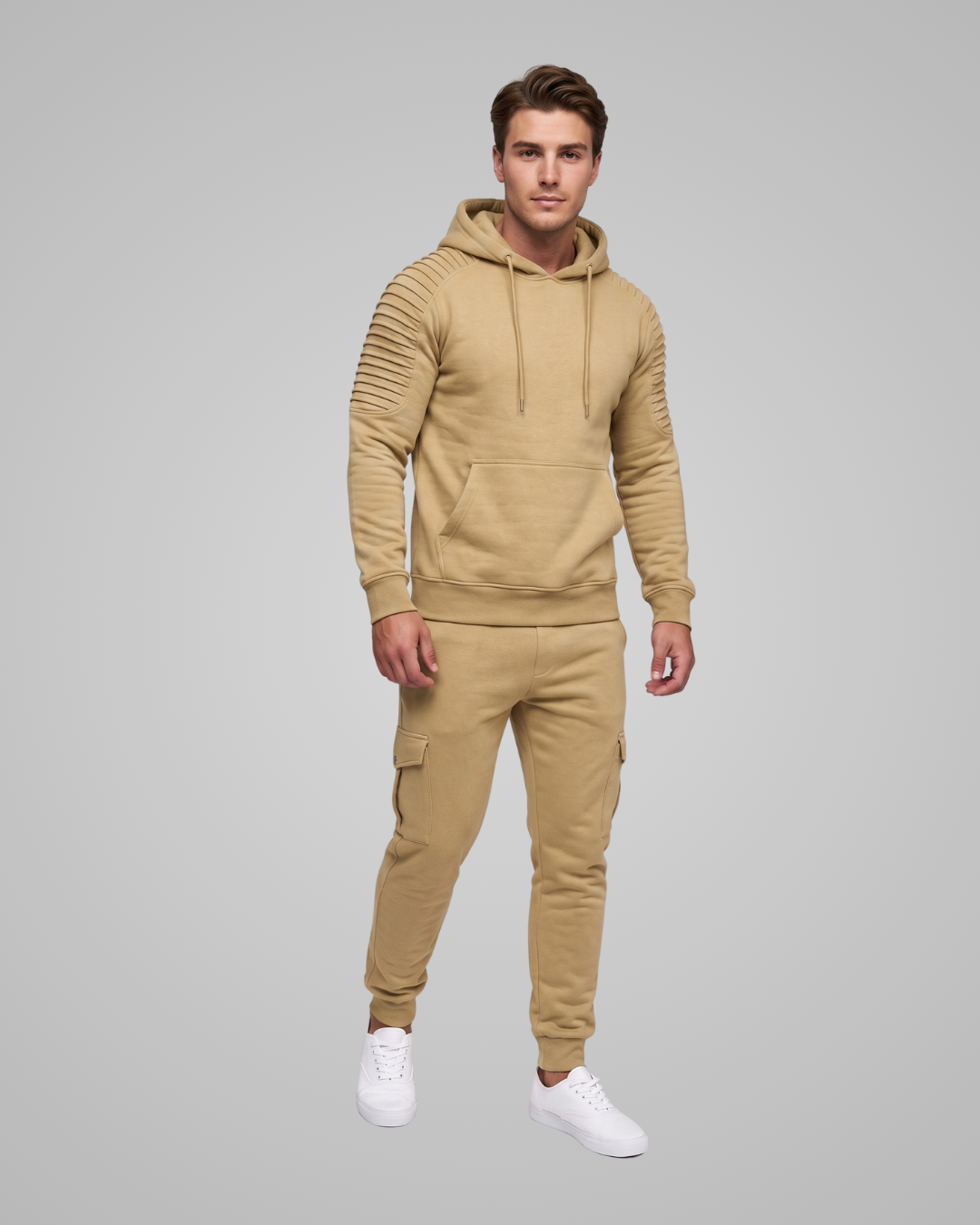 A man modeling a matching tan hoodie and jogger pants set with white sneakers, standing against a solid gray studio background.