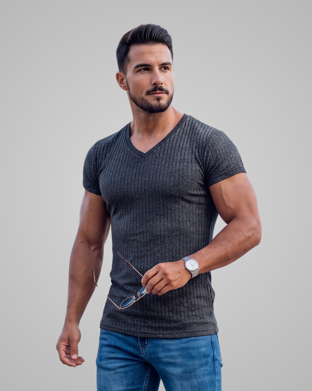 A muscular man wearing a dark gray ribbed v-neck t-shirt and blue jeans, holding a pair of eyeglasses against a solid light gray background.