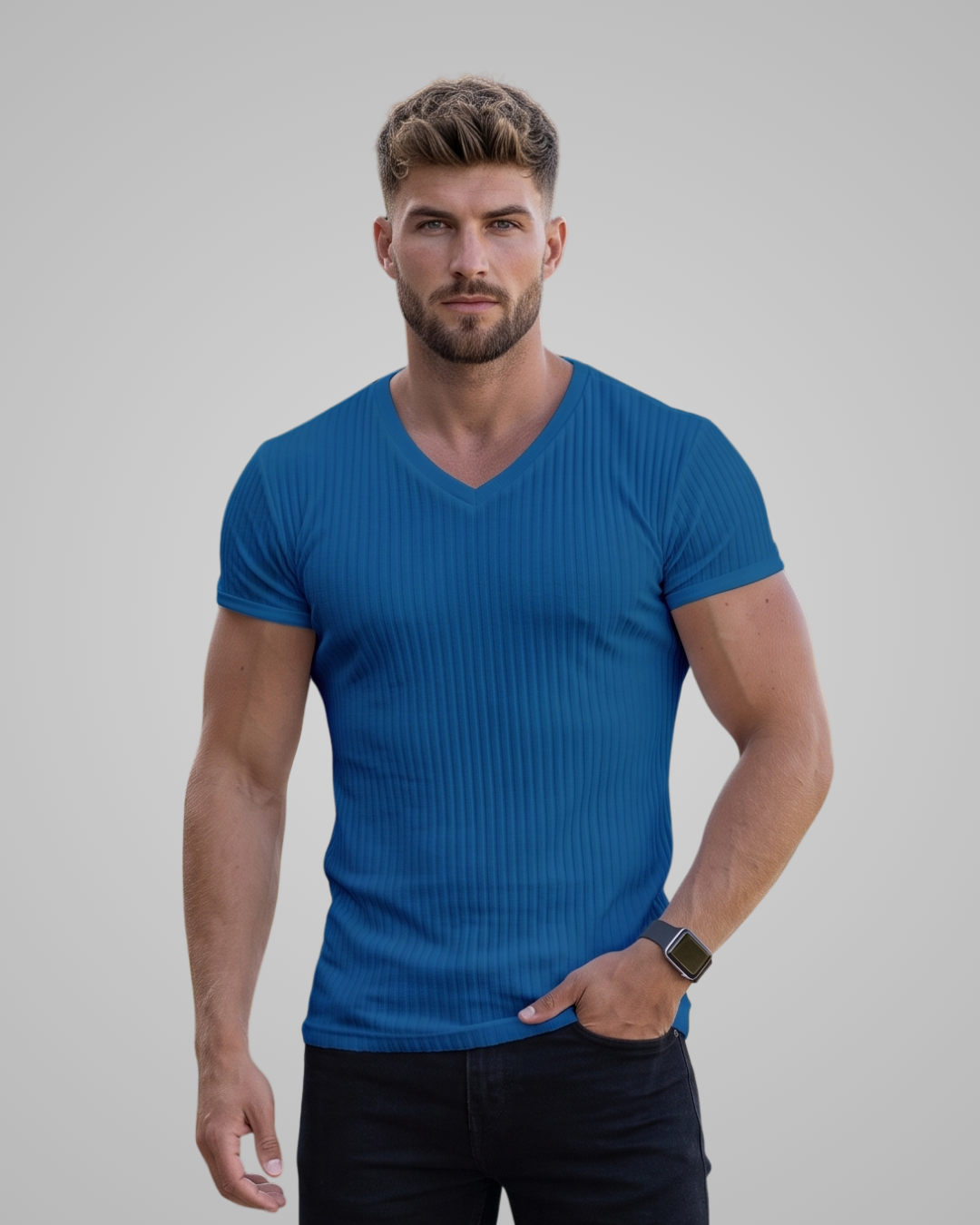 A muscular man wearing a blue ribbed short-sleeve v-neck t-shirt and dark jeans, standing against a solid gray studio background.