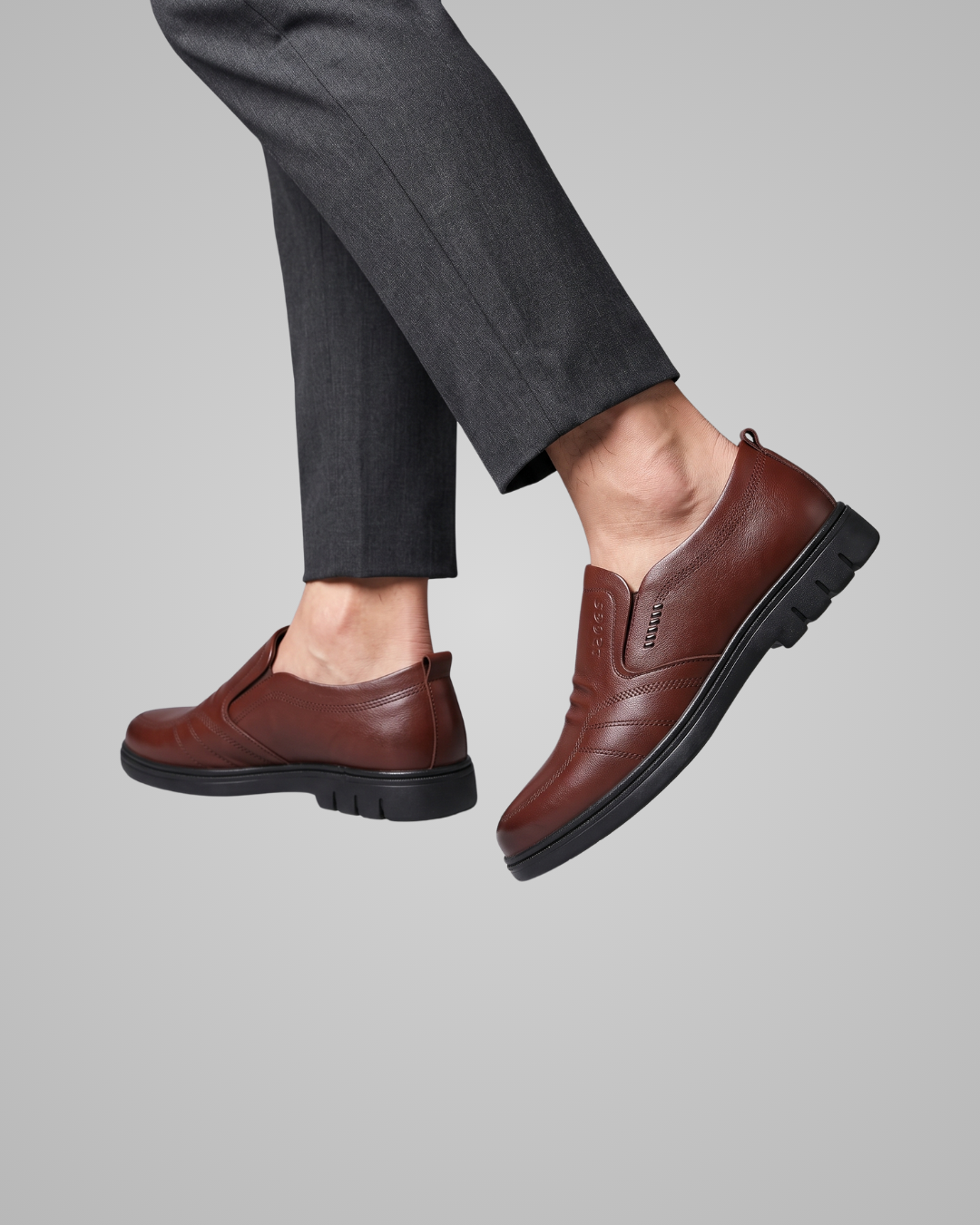 A pair of men's brown leather slip-on shoes with black soles, shown on a model wearing dark gray trousers against a solid gray background.